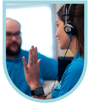 At the University of St. Augustine for Health Sciences Texas, a young woman wearing headphones signals with a raised hand to a man sitting across from her. Participating in SLP programs.