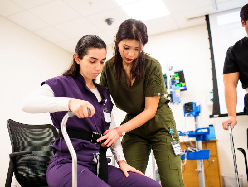 Two women in scrubs, one sitting and holding a cane while another helps secure a belt around her waist, in a clinical or training environment.