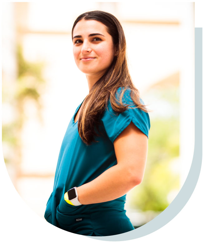 A young woman with long brown hair, wearing teal scrubs and a smartwatch, smiling at the camera.