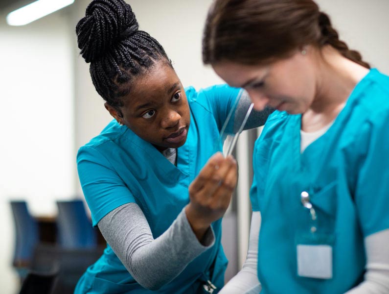 A medical professional in teal scrubs demonstrates a procedure to a colleague.