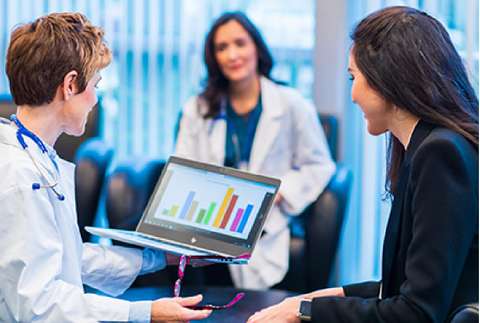 Three individuals in a healthcare setting, with one doctor presenting data on a laptop to a woman in a business suit, and another doctor observing.