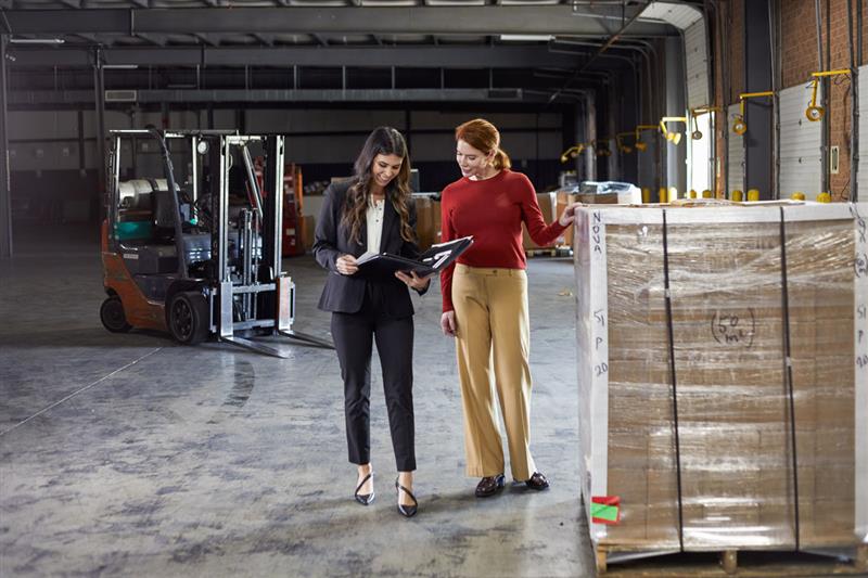 Two women discussing something while standing next to a large pallet of goods in a warehouse.