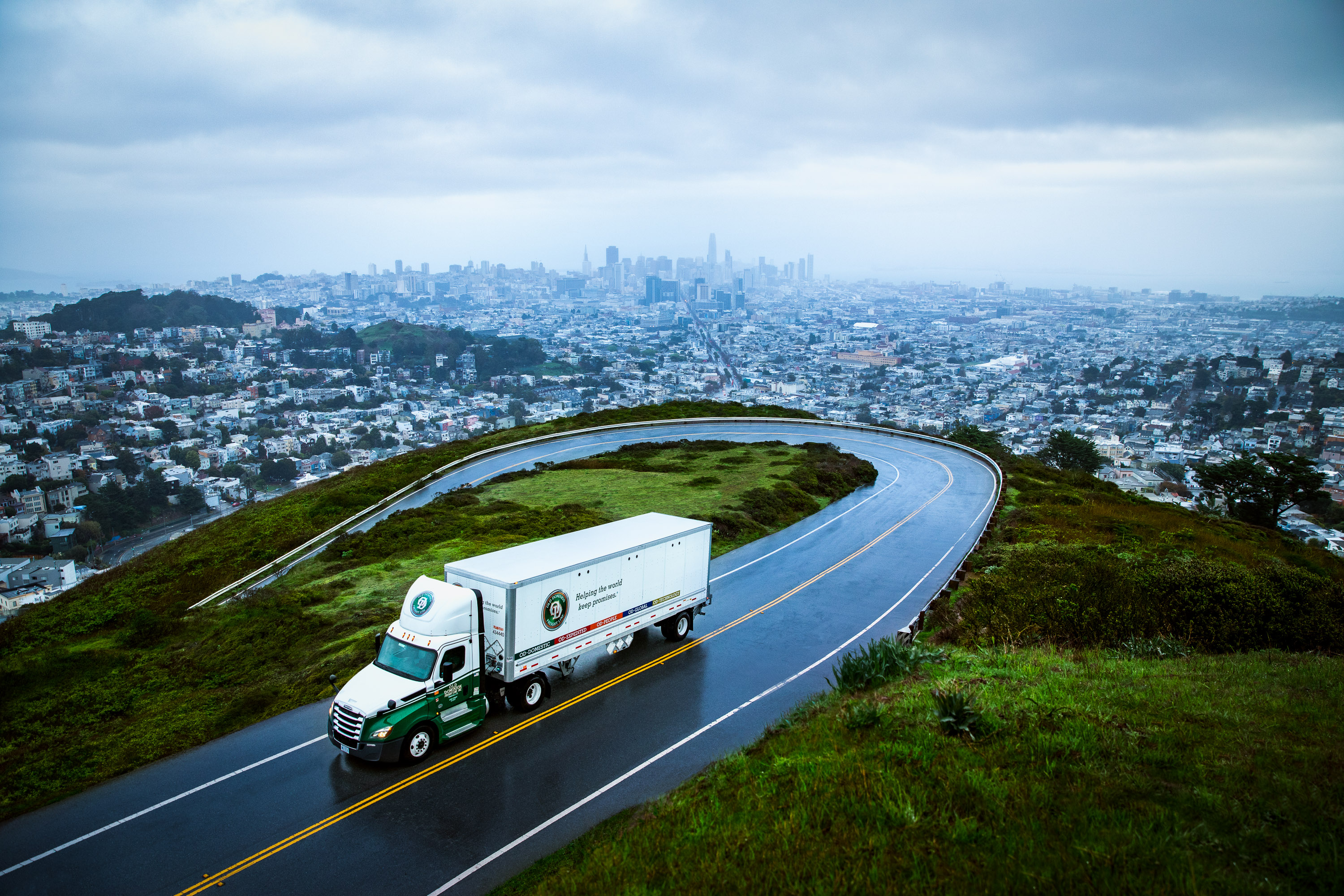 A white and green Old Dominion Freight Line truck drives on a winding road overlooking a city skyline under a cloudy sky.