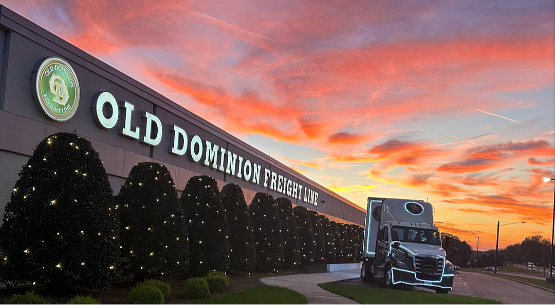 A semi-truck with glowing neon accents parked in front of an Old Dominion Freight Line building, with a vibrant orange and red sunset sky.