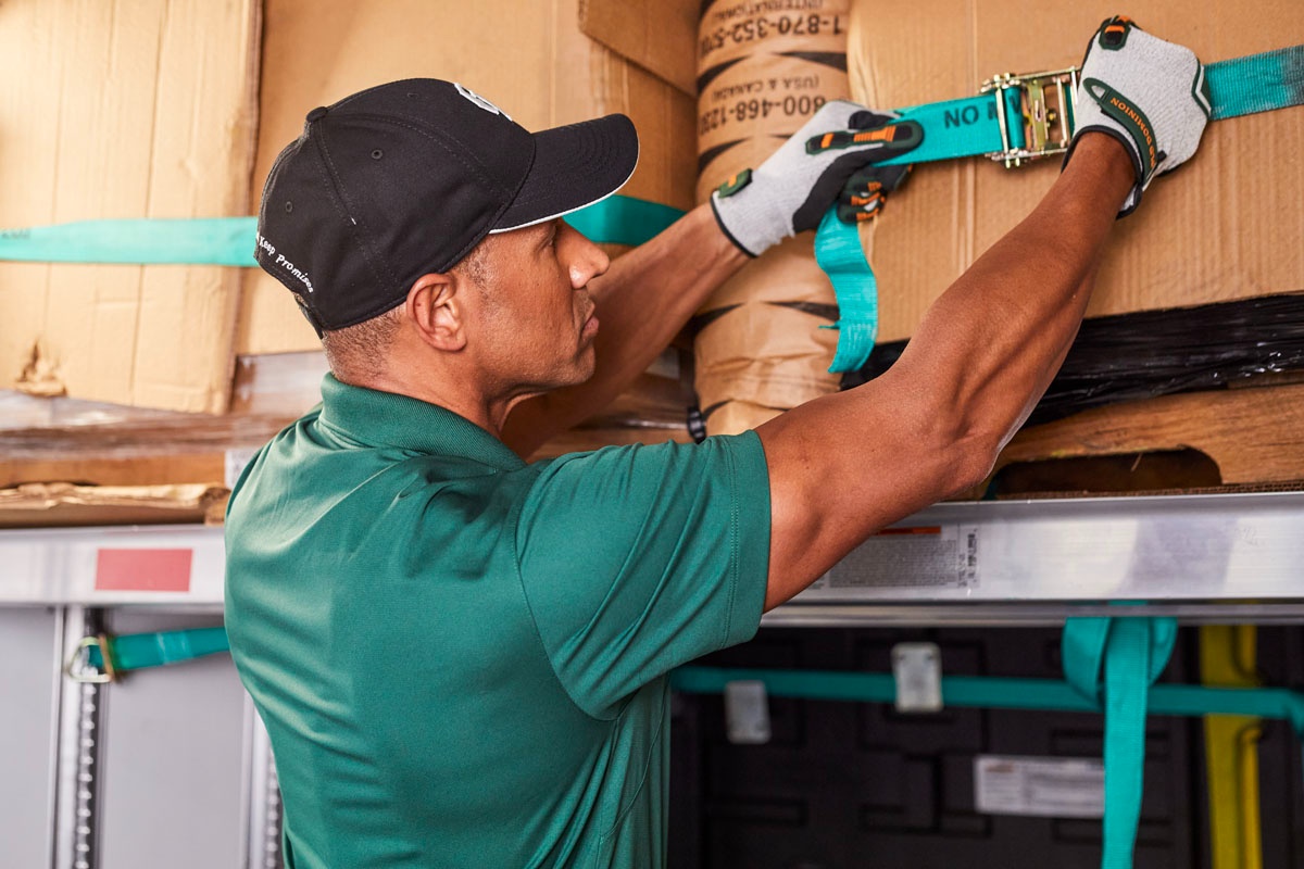 Close-up of a man wearing a black cap and work gloves, tightening a blue ratchet strap around stacked boxes.