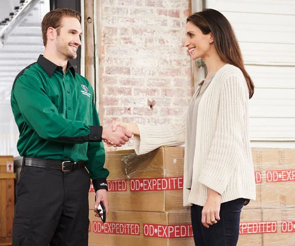 A delivery driver in a green uniform shakes hands with a smiling customer next to stacked boxes labeled 'EXPEDITED OD'.