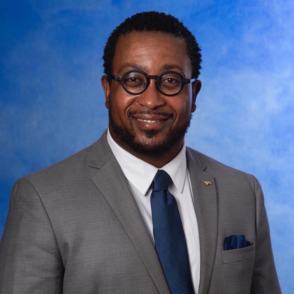 A smiling man in a grey suit, blue tie, and round glasses against a blue background.