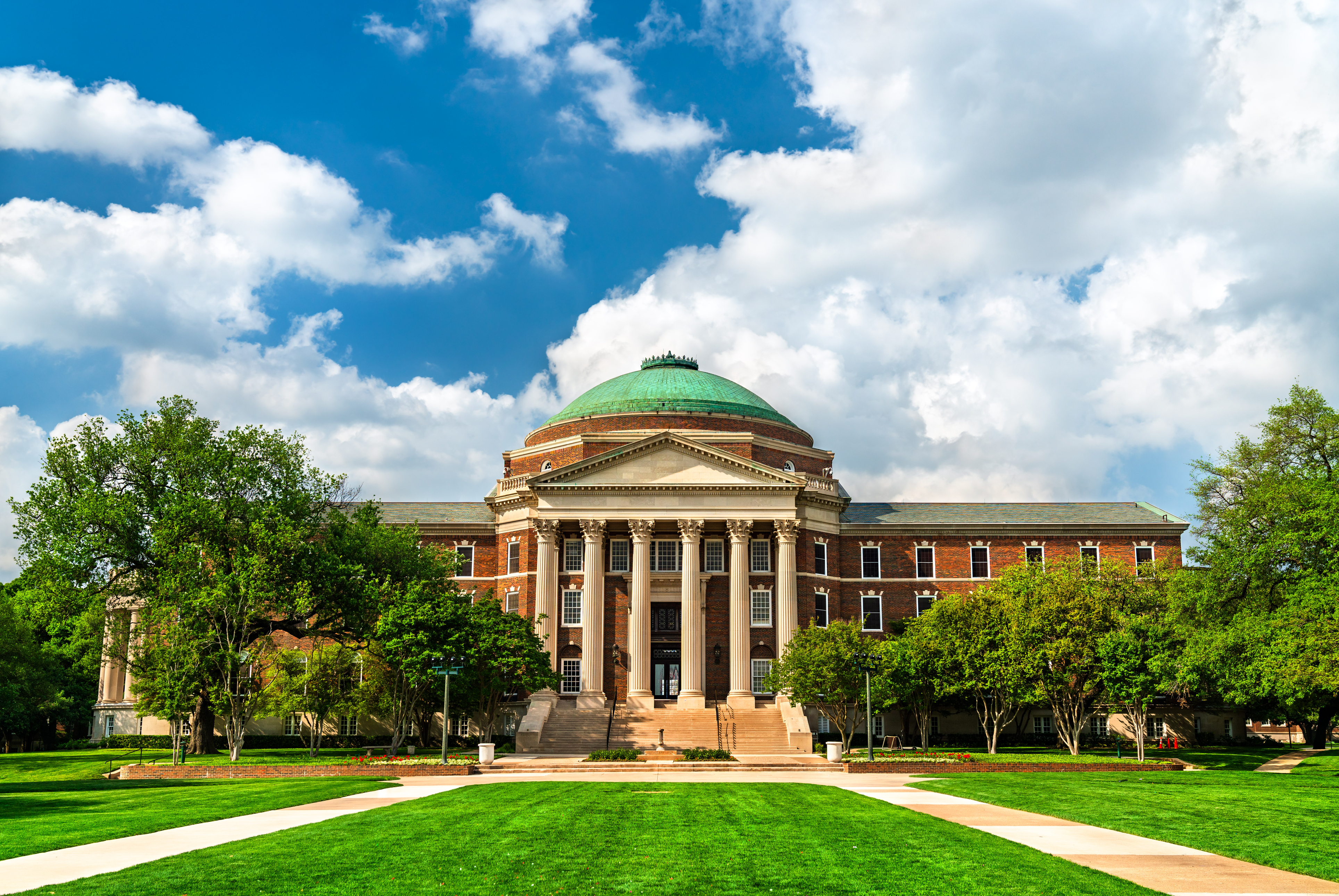 A grand, classical-style building with a green dome and large columns, set amidst lush green lawns and trees under a blue sky with white clouds.