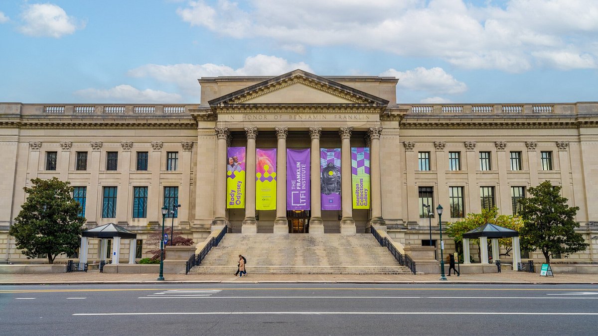 The Franklin Institute building, a grand neoclassical structure with columns, steps, and colorful banners advertising exhibits.