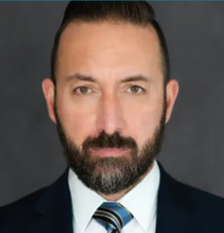Headshot of a man with a dark beard and mustache, wearing a navy suit and a striped blue and gold tie.