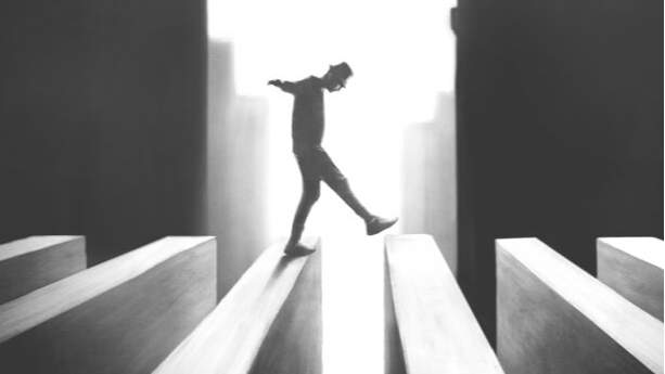 Article image - a black and white photo of a person walking on a precarious path of separated blocks, their arms outstretched for balance, against a stark white backdrop.