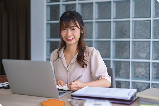 A young woman sits at a desk, working on a laptop and taking notes.