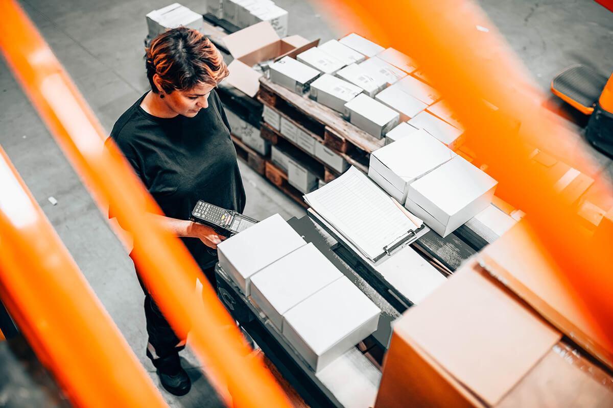 Warehouse worker scans packaged inventory beside stacked boxes and a clipboard, viewed through orange safety rails.