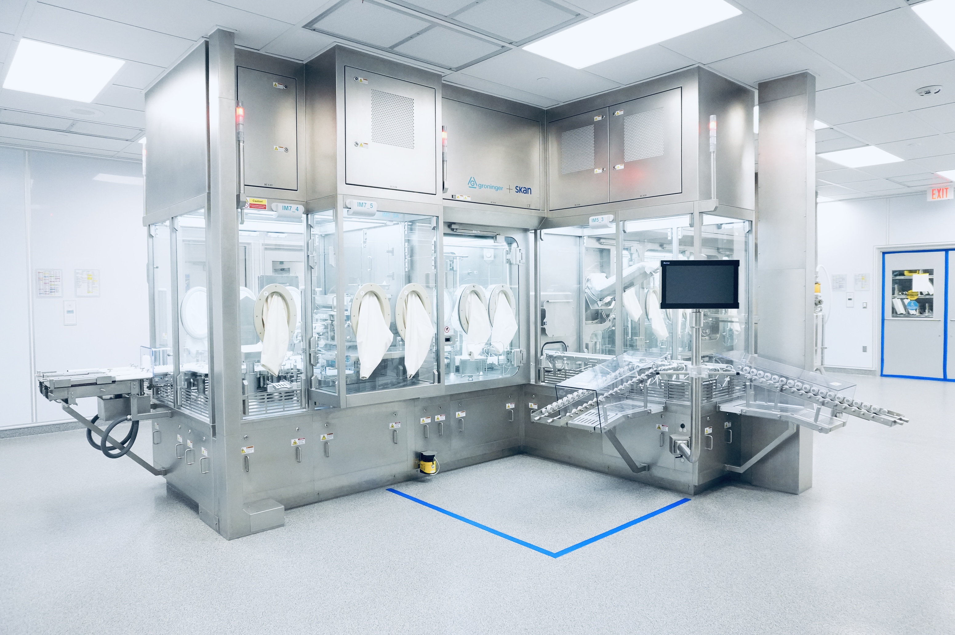 A modern cleanroom featuring an automated Groninger + Skan manufacturing line with glass enclosures, white gloves, and a system for conveying small containers.